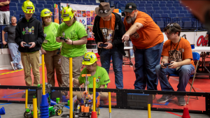 Two Mississippi teams, one from Bogue Chitto Attendance Center in green and another, from Wesson, in orange, compete in the Mississippi FIRST Tech Championship. Four students hold video-game controllers and two robots appear in the foreground.