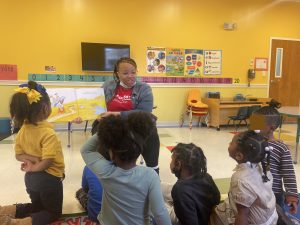 Mississippi Jumpstart volunteer reads to a group of preschoolers. The room is yellow and covered in educational posters. Six children gather around the reader.
