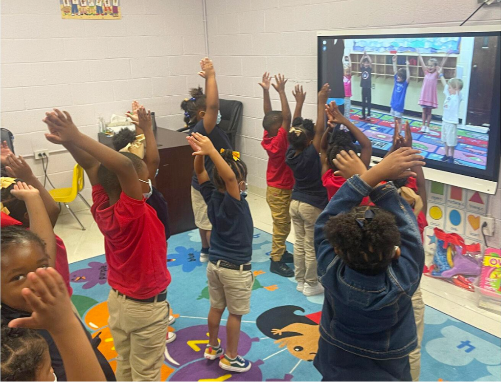 A classroom of children raise their arms and do yoga alongside children on tv who demonstrate different moves.
